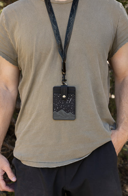 A tight close-up shot focusing on the front of the Midnight Mountain vertical card holder, highlighting the large clear plastic viewing window and meticulous white stitching. The clean black and white starry sky landscape design surrounds the badge sleeve, creating a professional look for school or work access cards.