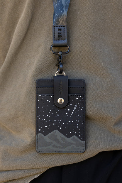 An overhead view of the Midnight Mountain ID badge holder and matching neck lanyard laid flat on a rustic dark wood log. The product displays a striking graphic design of dark gray mountain ridges under a black night sky covered in white stars, complete with an antique bronze metal attachment clip.