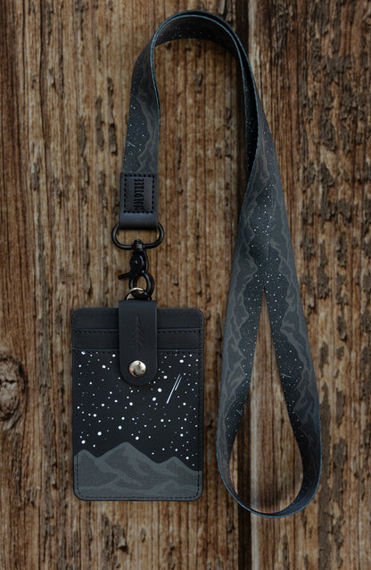 A close-up lifestyle shot highlighting the back of the Midnight Mountain ID badge holder being worn outdoors on a neck lanyard. The photo showcases the premium texture of the black synthetic vegan leather card slots and the secure brown button strap with a debossed pine tree icon that safely protects credit cards or cash.