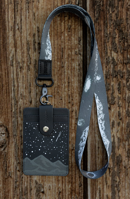 A waist-up lifestyle view of a person outdoors wearing the black and gray Midnight Mountain ID badge holder clipped to a matching mountain-themed neck lanyard. This durable vegan leather vertical badge wallet hangs neatly against a black t-shirt, showing how teachers, nurses, and office workers can wear it comfortably on the job.
