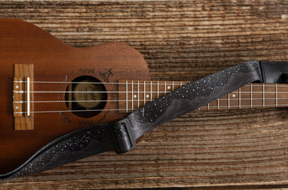 Black Ukulele Strap Strap Printed with Mountains and Stars lying across Ukulele on wood floor