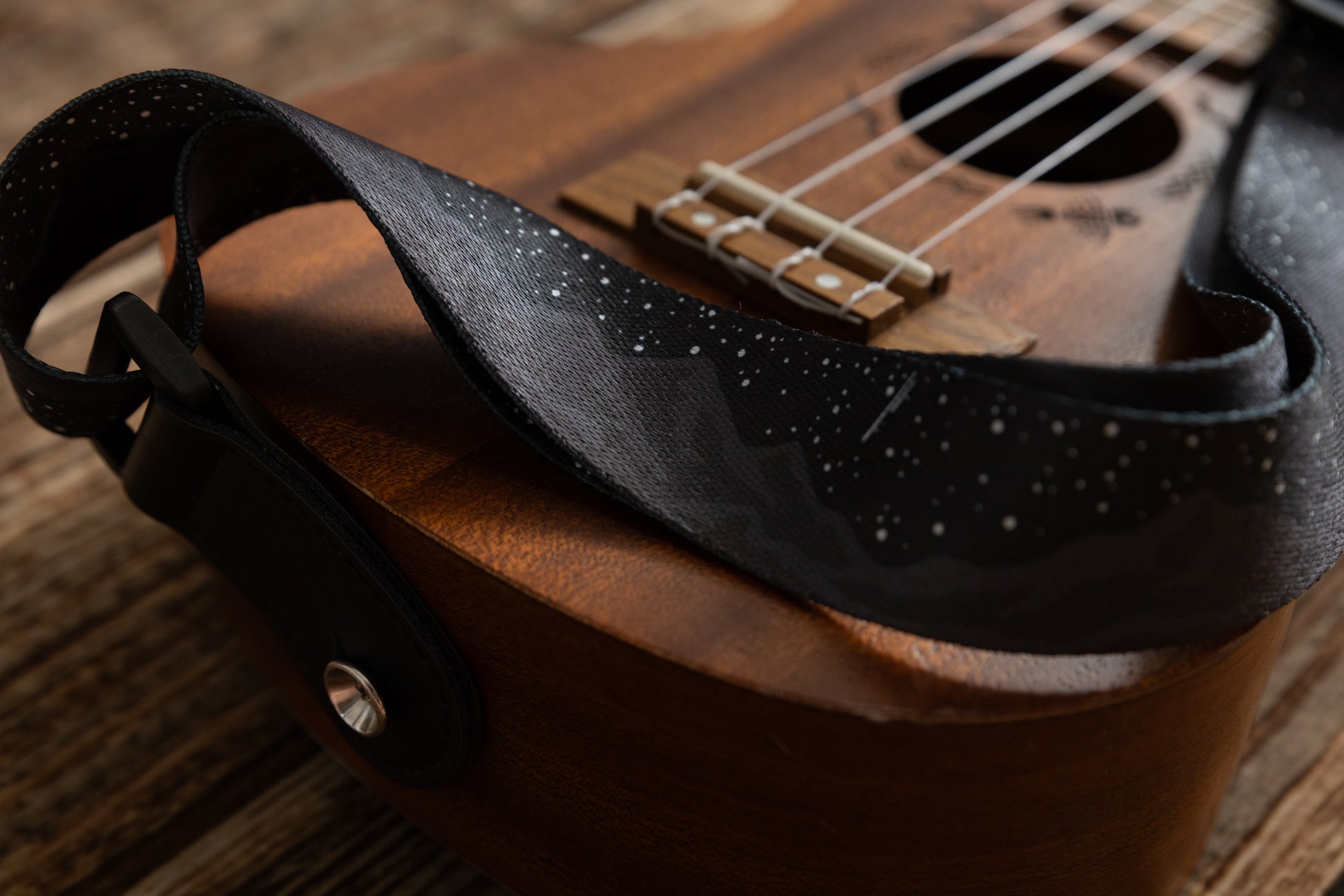 Black Ukulele Strap Strap Printed with Mountains and Stars lying across Ukulele on wood floor