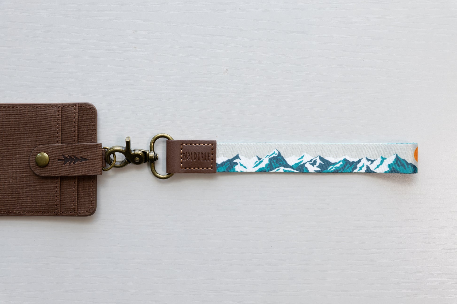 The Mountain Horizon wristlet keychain attached to a brown vegan-leather snap wallet on a white background, showing the soft gray strap printed with a navy and teal mountain silhouette and orange sun accent.