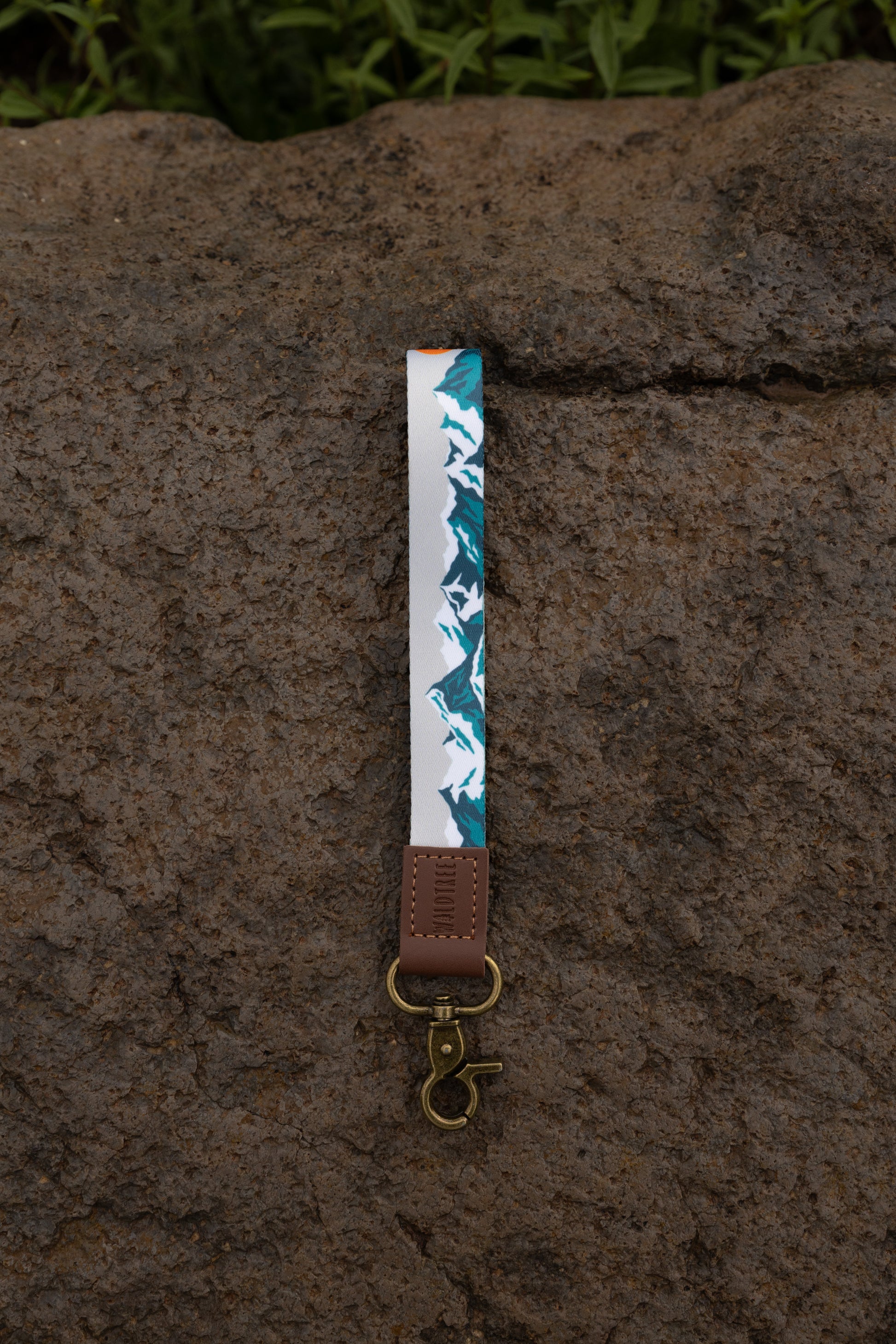 The Mountain Horizon wristlet keychain hanging against a dark stone wall backdrop, showing the soft gray strap printed with a navy and teal mountain silhouette, the brown leather WILDTREE tab, and the bronze swivel clasp.