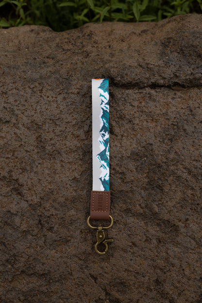 The Mountain Horizon wristlet keychain hanging against a dark stone wall backdrop, showing the soft gray strap printed with a navy and teal mountain silhouette, the brown leather WILDTREE tab, and the bronze swivel clasp.