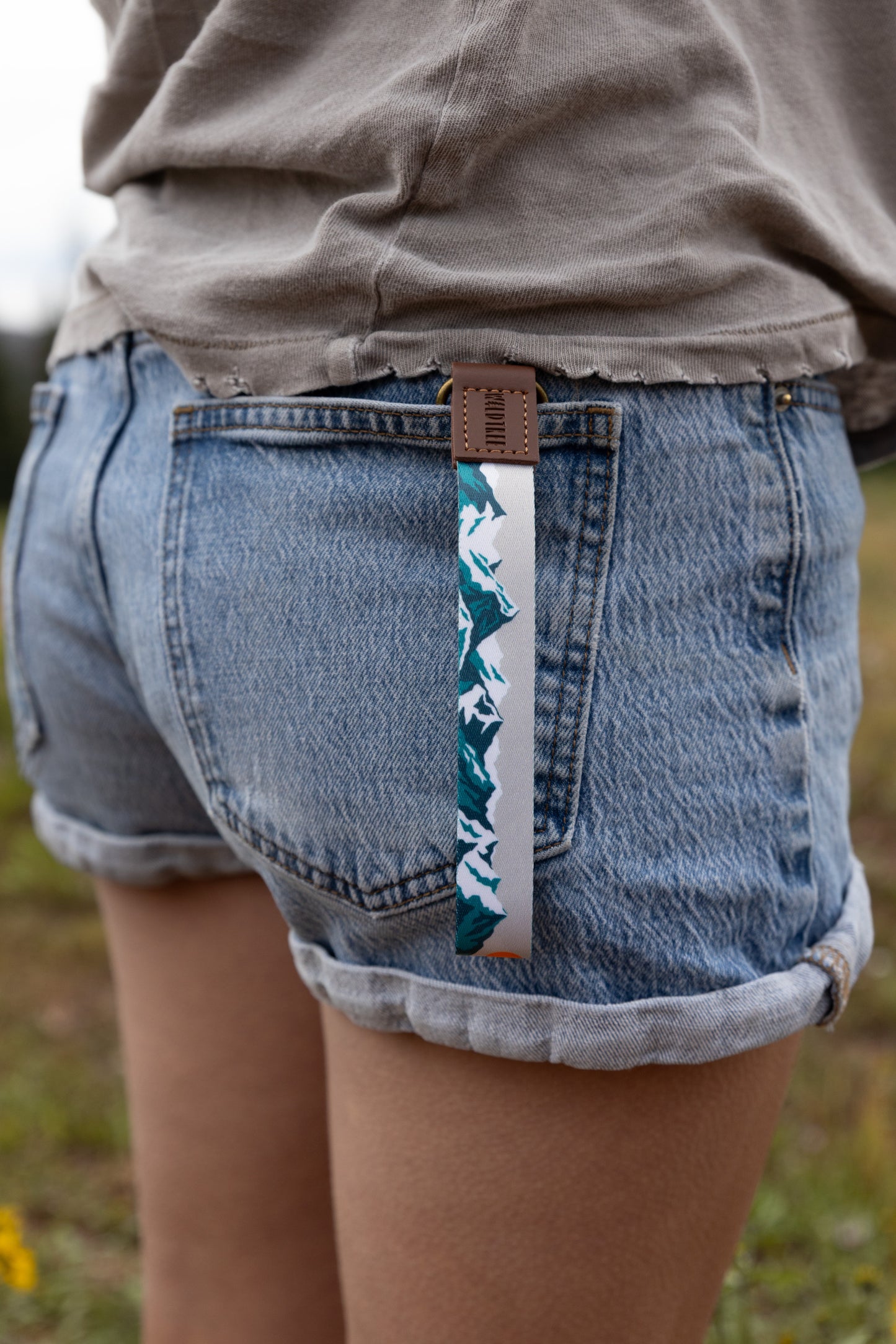 The Mountain Horizon wristlet keychain clipped to the back belt loop of denim shorts, the soft gray strap printed with a navy and teal mountain silhouette hanging against the wearer's leg.