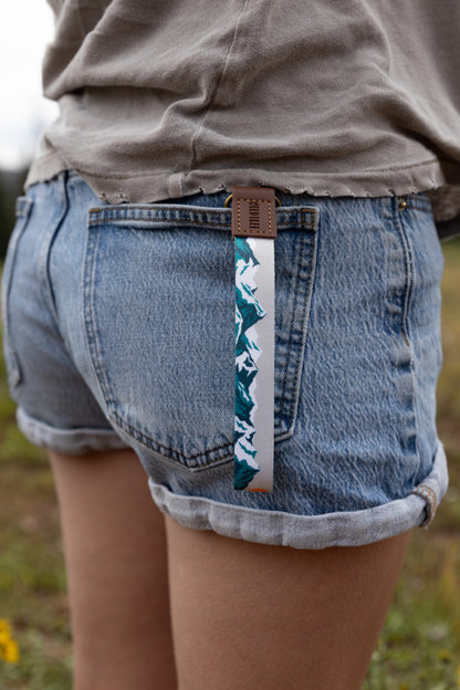 The Mountain Horizon wristlet keychain clipped to the back belt loop of denim shorts, the soft gray strap printed with a navy and teal mountain silhouette hanging against the wearer's leg.