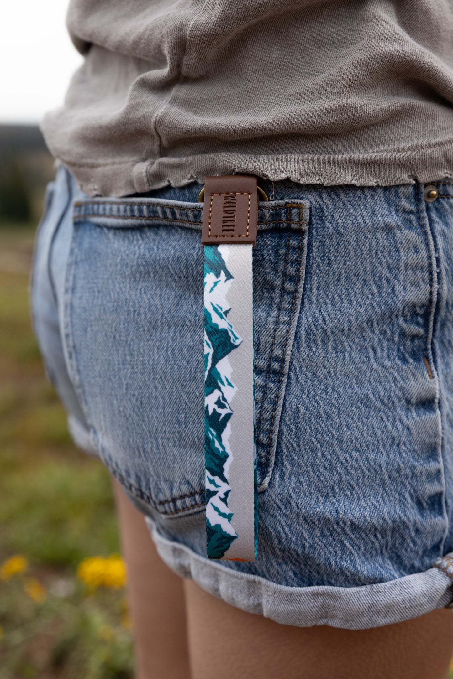 Close-up from above of the Mountain Horizon wristlet keychain clipped to a denim shorts belt loop, the soft gray strap printed with a navy and teal mountain silhouette hanging against the wearer's leg.