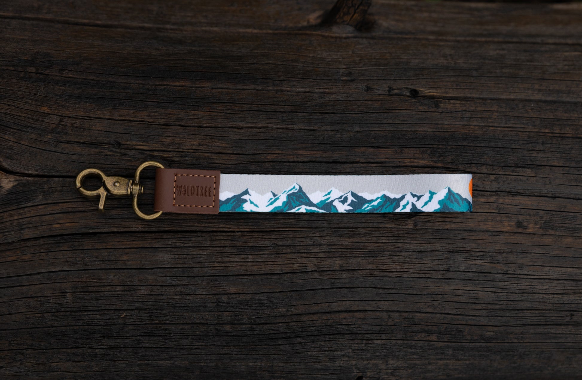 The Mountain Horizon wristlet keychain laid flat on a dark wood plank surface, showing the soft gray strap printed with a navy and teal mountain silhouette and orange sun accent, the brown leather WILDTREE tab, and the bronze swivel clasp.
