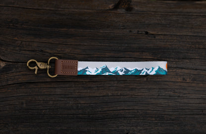 The Mountain Horizon wristlet keychain laid flat on a dark wood plank surface, showing the soft gray strap printed with a navy and teal mountain silhouette and orange sun accent, the brown leather WILDTREE tab, and the bronze swivel clasp.