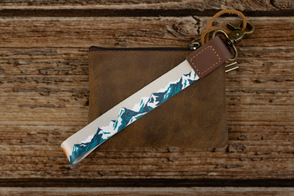 The Mountain Horizon wristlet keychain attached to a set of keys and draped across a brown leather zip pouch on a wood surface, showing the soft gray strap printed with a navy and teal mountain silhouette.