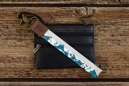 The Mountain Horizon wristlet keychain attached to a set of keys and draped across a black leather zip pouch on a wood surface, showing the soft gray strap printed with a navy and teal mountain silhouette.
