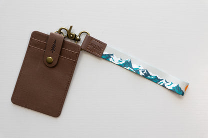 The Mountain Horizon wristlet keychain attached to a brown vegan-leather card holder on a white background, showing the soft gray strap printed with a navy and teal mountain silhouette, the brown leather WILDTREE tab, and bronze clasps.