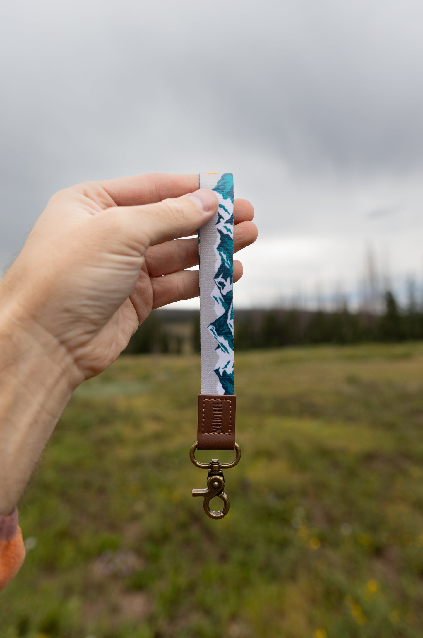 A hand holds the Mountain Horizon wristlet keychain up against a cloudy sky and grassy field, showing the soft gray strap printed with a navy and teal mountain silhouette, the brown leather WILDTREE tab, and bronze clasps.