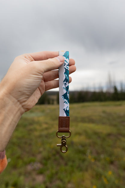 A hand holds the Mountain Horizon wristlet keychain up against a cloudy sky and grassy field, showing the soft gray strap printed with a navy and teal mountain silhouette, the brown leather WILDTREE tab, and bronze clasps.