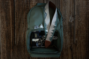 Wildtree Mountain and tree Camera strap featuring green trees and blue mountain sitting on green backpack -best camera strap for canon cameras