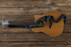 Black guitar strap with trees and stars attached to guitar lying on wood floor
