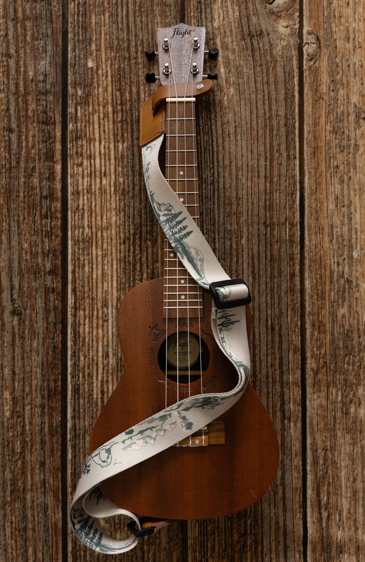 National Park-Themed Ukulele Strap printed with designs of 12 popular national parks along the length of the strap. connected to Ukulele lying on wood floor