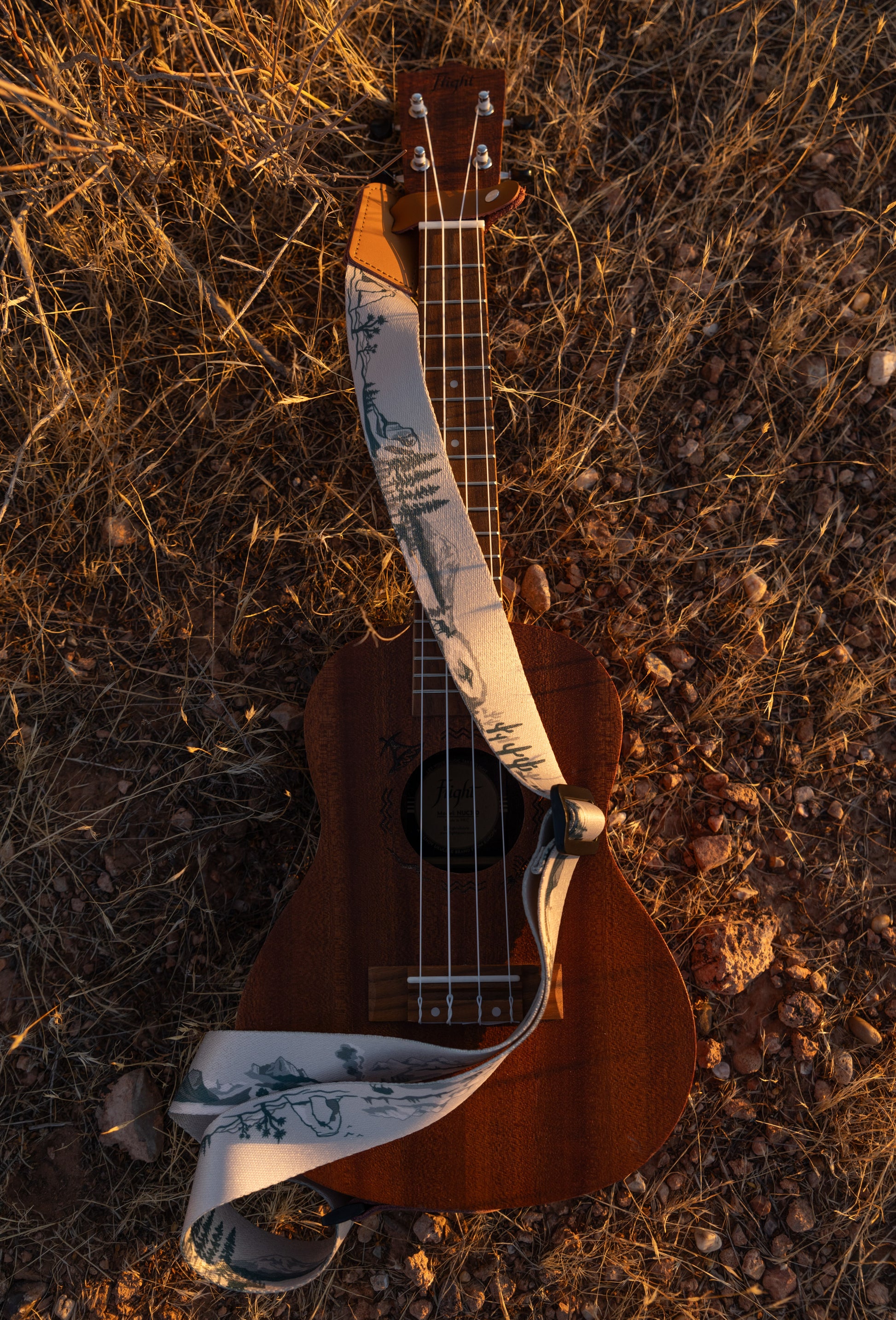 National Park-Themed Ukulele Strap printed with designs of 12 popular national parks along the length of the strap. connected to Ukulele lying on desert ground