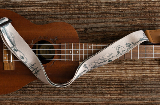 National Park-Themed Ukulele Strap printed with designs of 12 popular national parks along the length of the strap. connected to Ukulele lying on wood floor