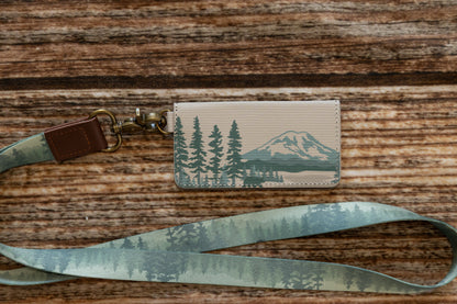 The SD card holder attached to a matching forest-patterned lanyard via a brass D-ring and swivel clasp, laid out on a wooden background.