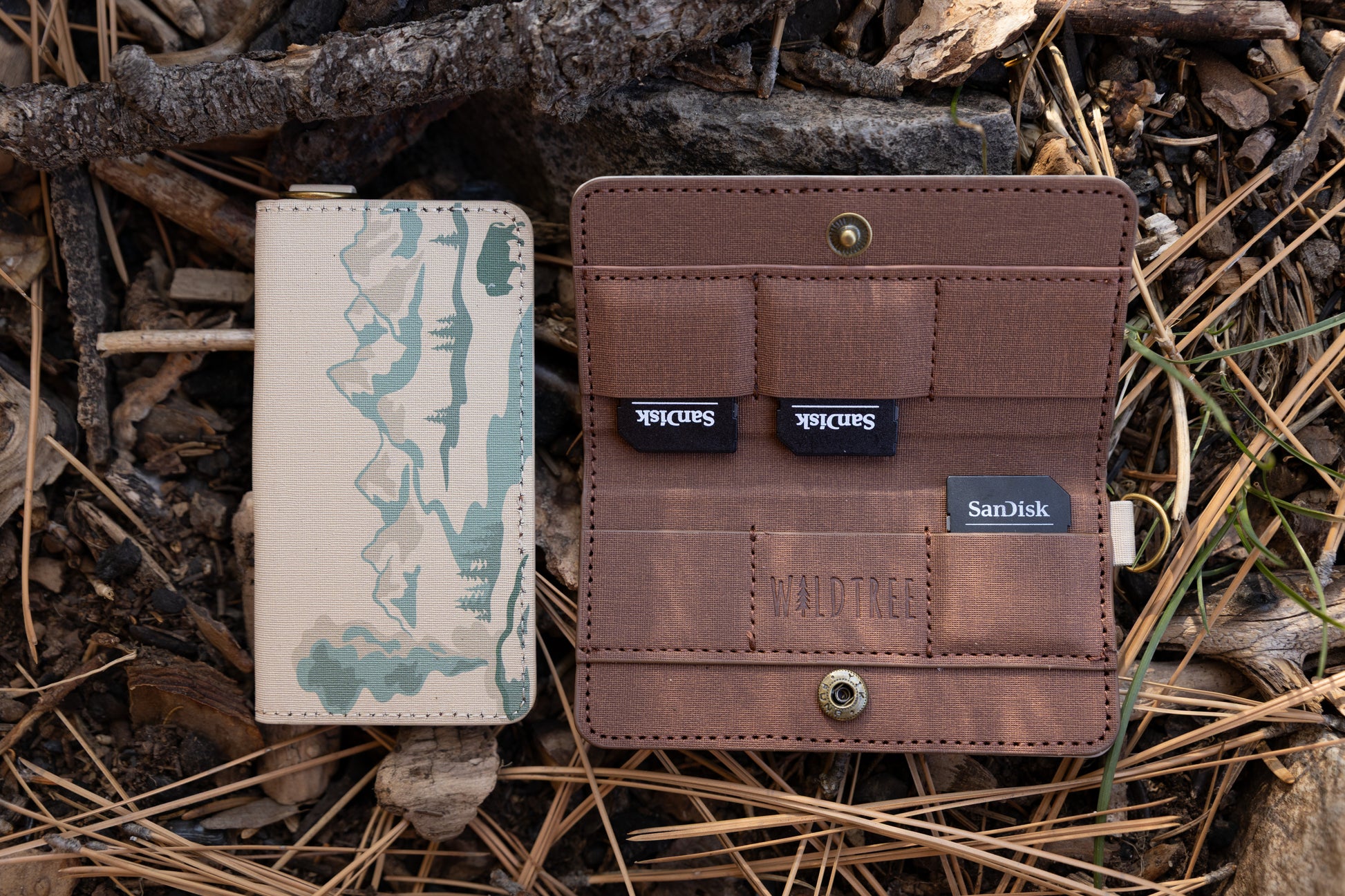 A side-by-side view of a tan fabric SD card holder. The left shows the closed front featuring a teal mountain and forest illustration; the right shows the open interior with brown pockets holding three SanDisk SD cards, featuring the "WILDTREE" logo.