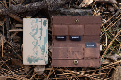 A side-by-side view of a tan fabric SD card holder. The left shows the closed front featuring a teal mountain and forest illustration; the right shows the open interior with brown pockets holding three SanDisk SD cards, featuring the "WILDTREE" logo.