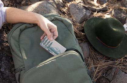 A lifestyle shot of a person placing the tan mountain-patterned SD card holder into the front pocket of a green backpack outdoors, with a green felt hat resting on the ground nearby.