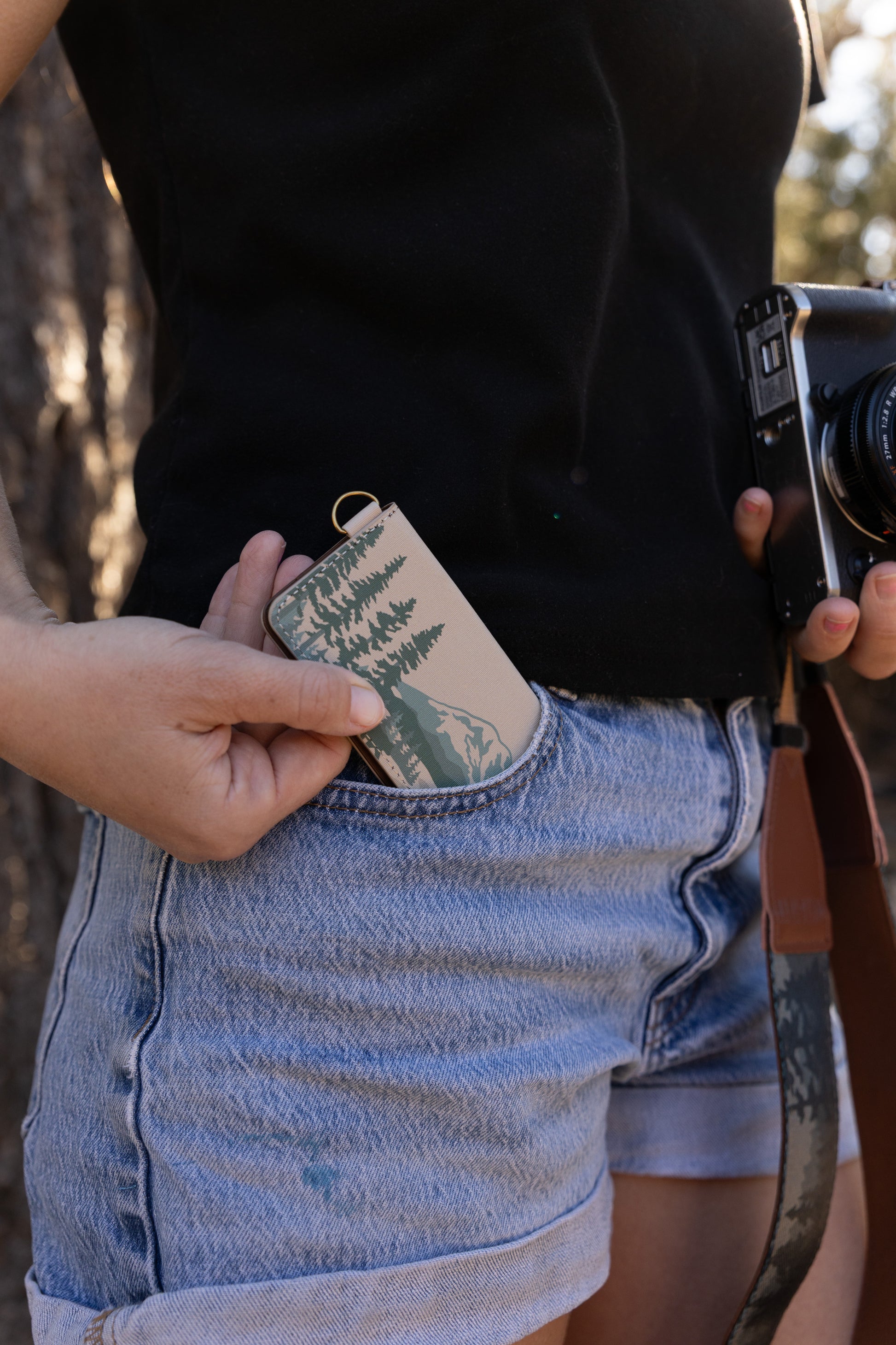 A close-up shot of a person wearing denim shorts and a black t-shirt, sliding the closed mountain-themed SD card holder into their front pocket while holding a camera.