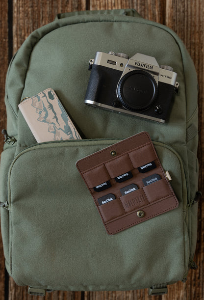 An overhead flat lay on a green backpack. A Fujifilm X-T30 camera sits at the top, while an open tan SD card holder filled with six SanDisk cards rests on the front pocket.