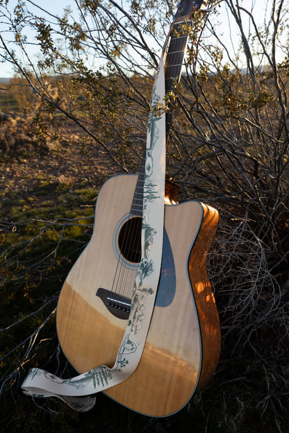 An acoustic-electric guitar leaning against a desert bush, showcasing the full length of the cream and green National Park strap draped over the body. The strap features distinct illustrations of cacti, mountains, and forests.