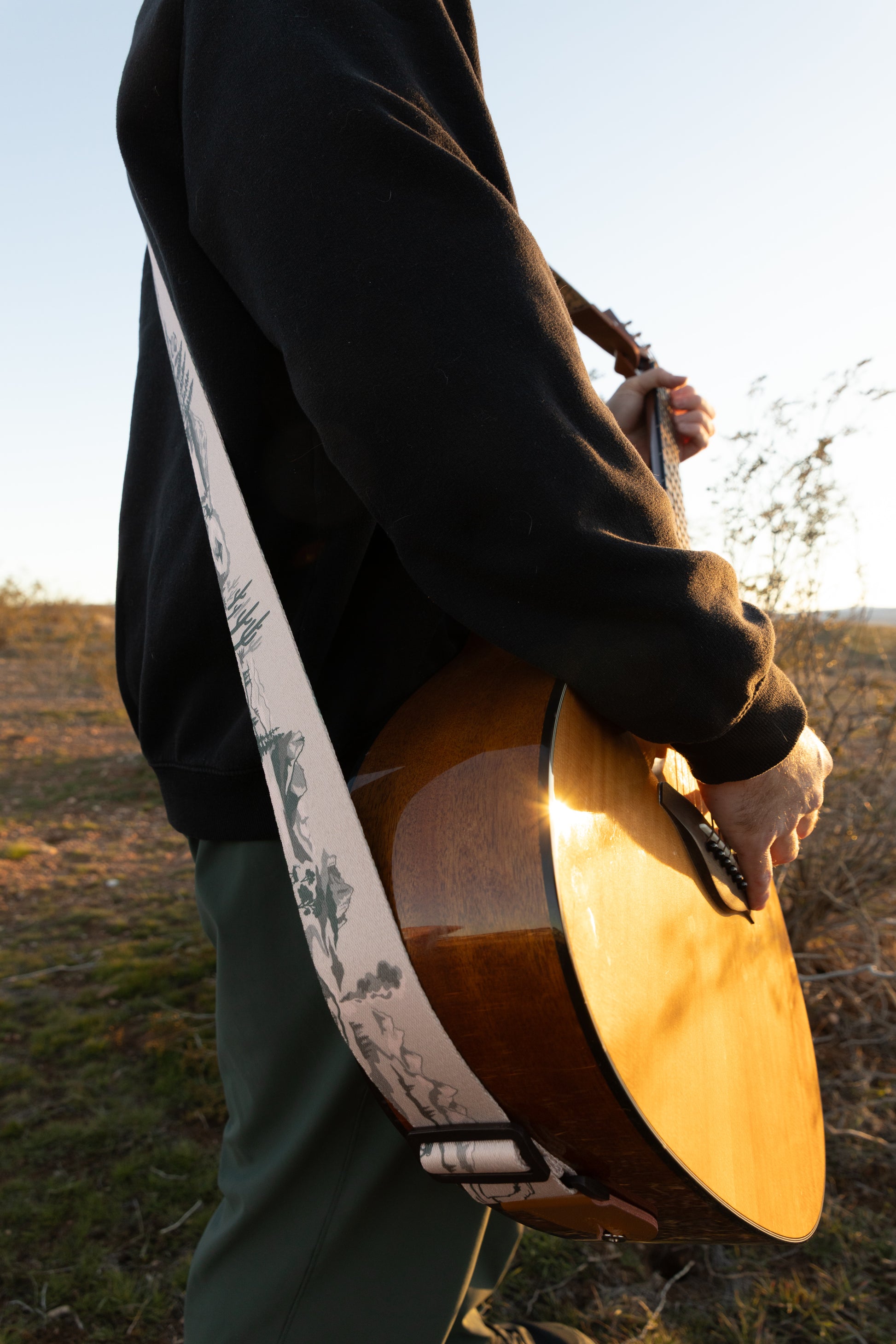 A side-profile view of a person standing in a desert landscape at sunset, wearing a National Park themed guitar strap. The low sun illuminates the side of the acoustic guitar, highlighting the detailed tree and mountain sketches on the strap.