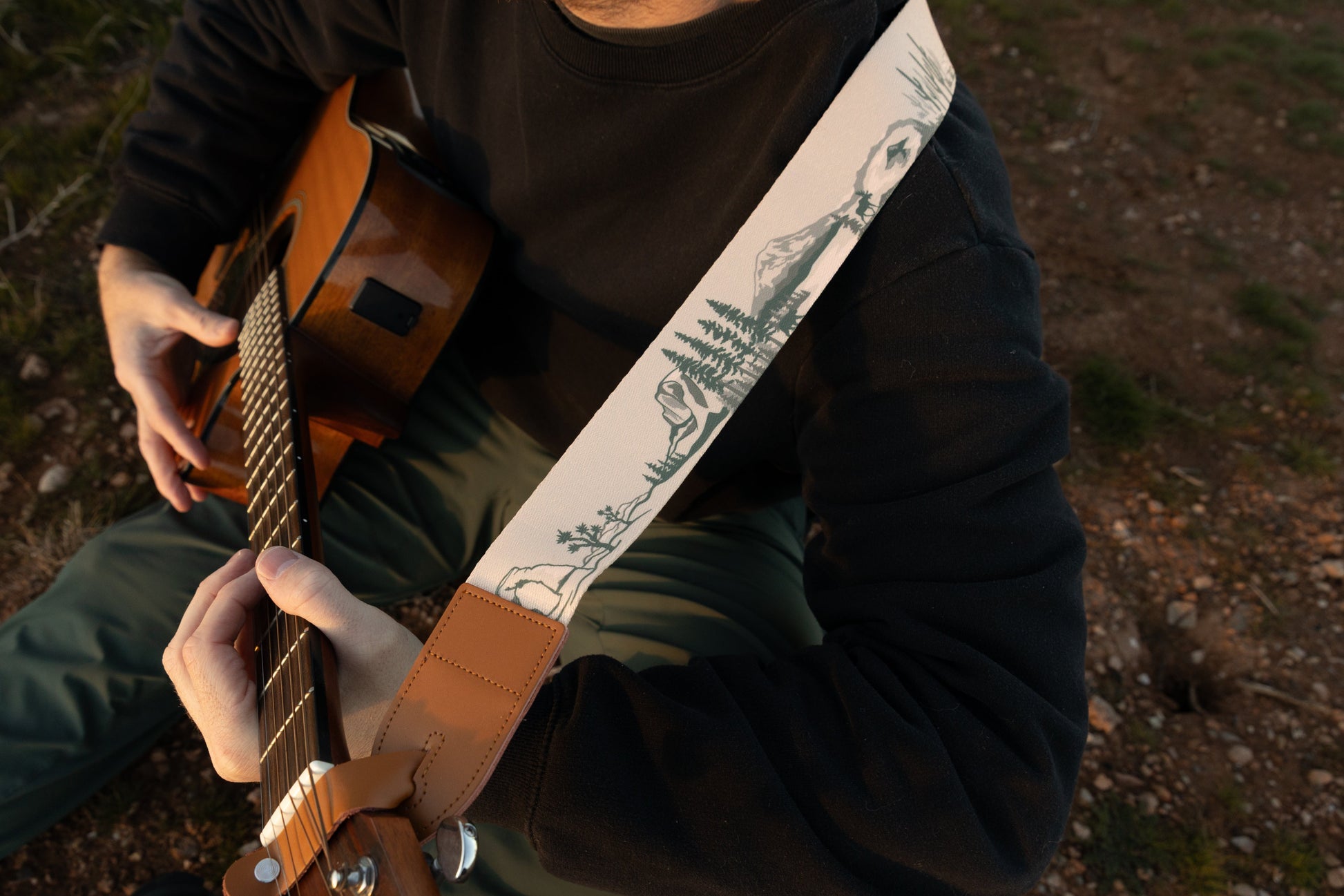 A close-up, high-angle view of a musician sitting outdoors, wearing a cream-colored guitar strap with green National Park illustrations. The strap features a tan leather end piece and is attached to a natural wood acoustic guitar.
