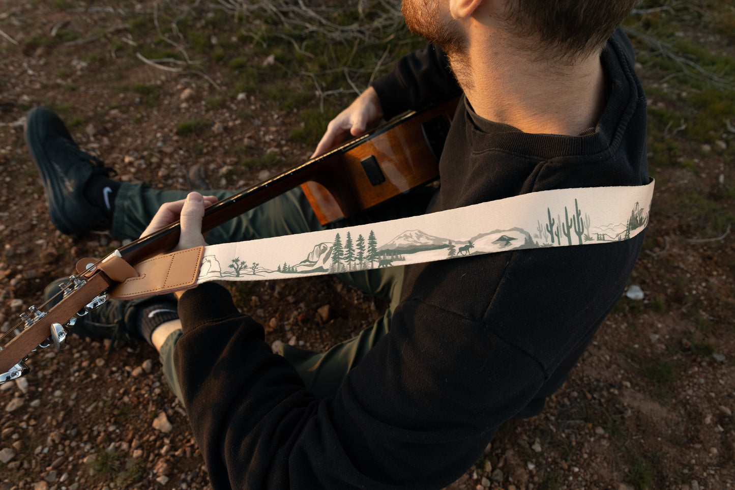 An over-the-shoulder view of a musician playing a guitar outdoors. The strap is pulled taut, providing a clear, horizontal view of the printed icons like the Joshua Tree, moose, and mountain ranges.