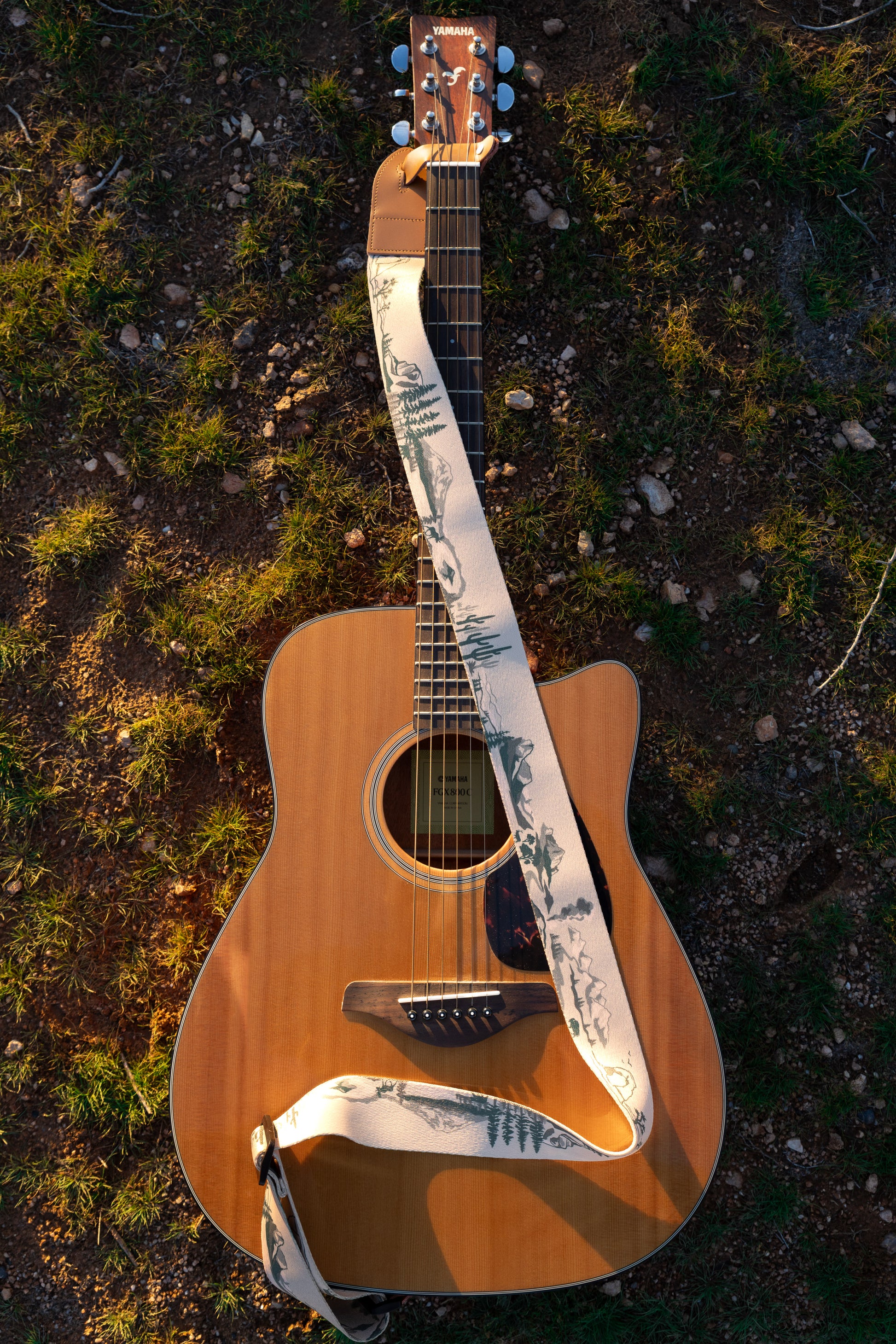 A top-down flat-lay image of an acoustic guitar resting on the ground. The National Park guitar strap is artfully coiled across the body, clearly showing the 12 different park illustrations including Saguaro cacti and Yosemite peaks.