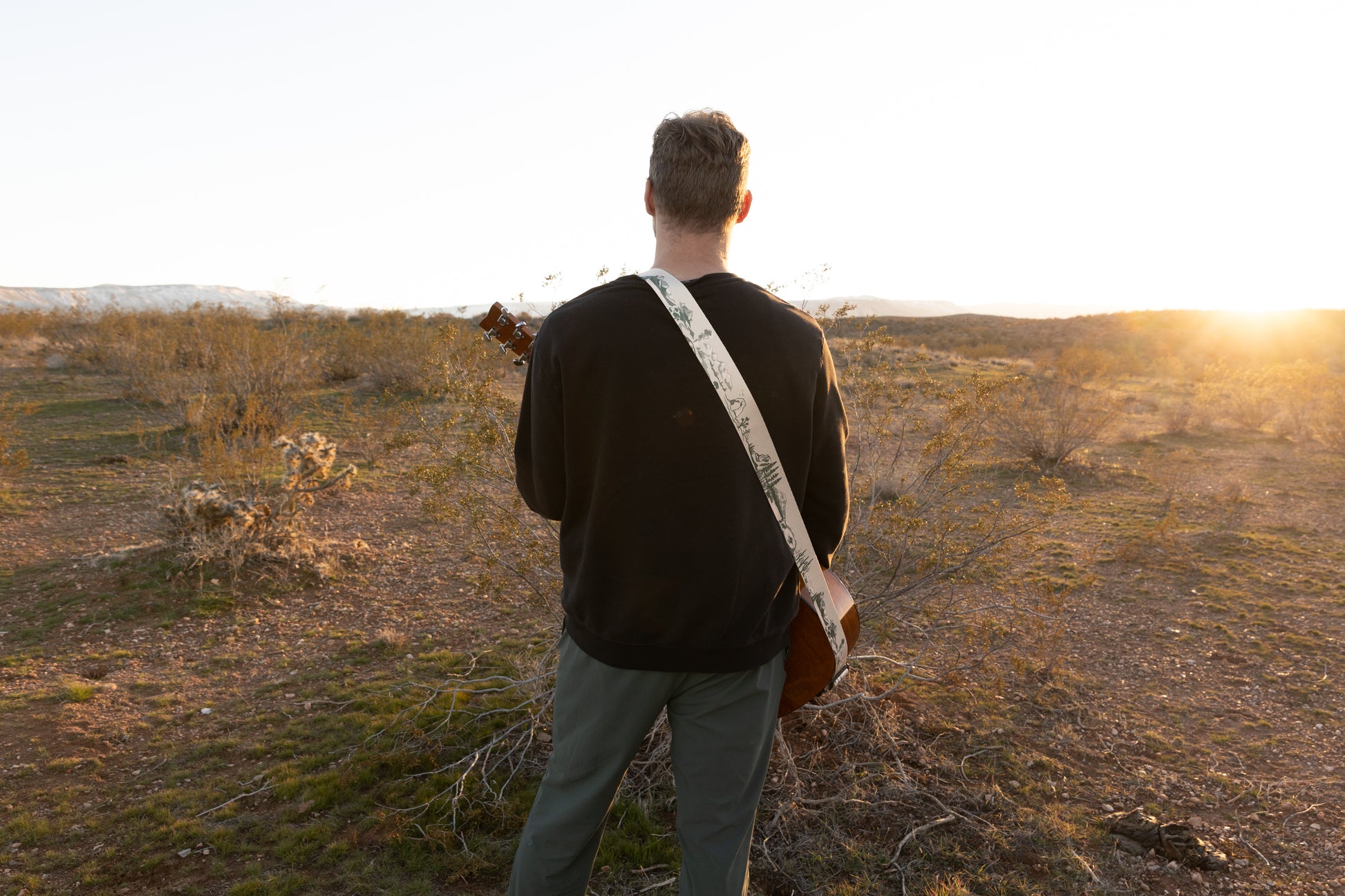 A wide, rear-view shot of a person standing in an open desert valley during golden hour, wearing a guitar across their back. The white and green National Park guitar strap stands out against a dark sweatshirt, showing the continuous landscape design.