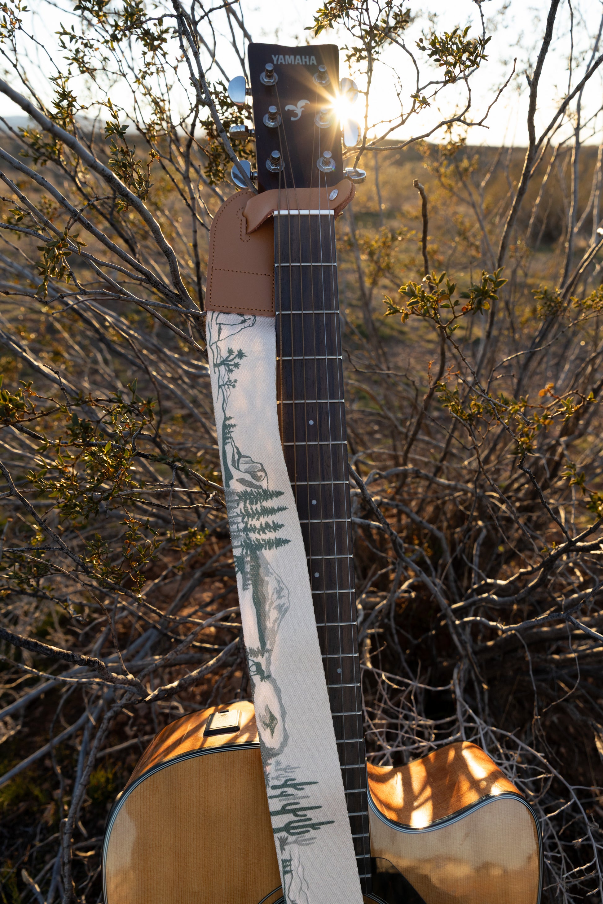 A detailed vertical shot of a Yamaha guitar headstock leaning against desert brush, with the sun peeking through the branches. The tan leather connector and the illustrated National Park strap are prominently displayed in the foreground.