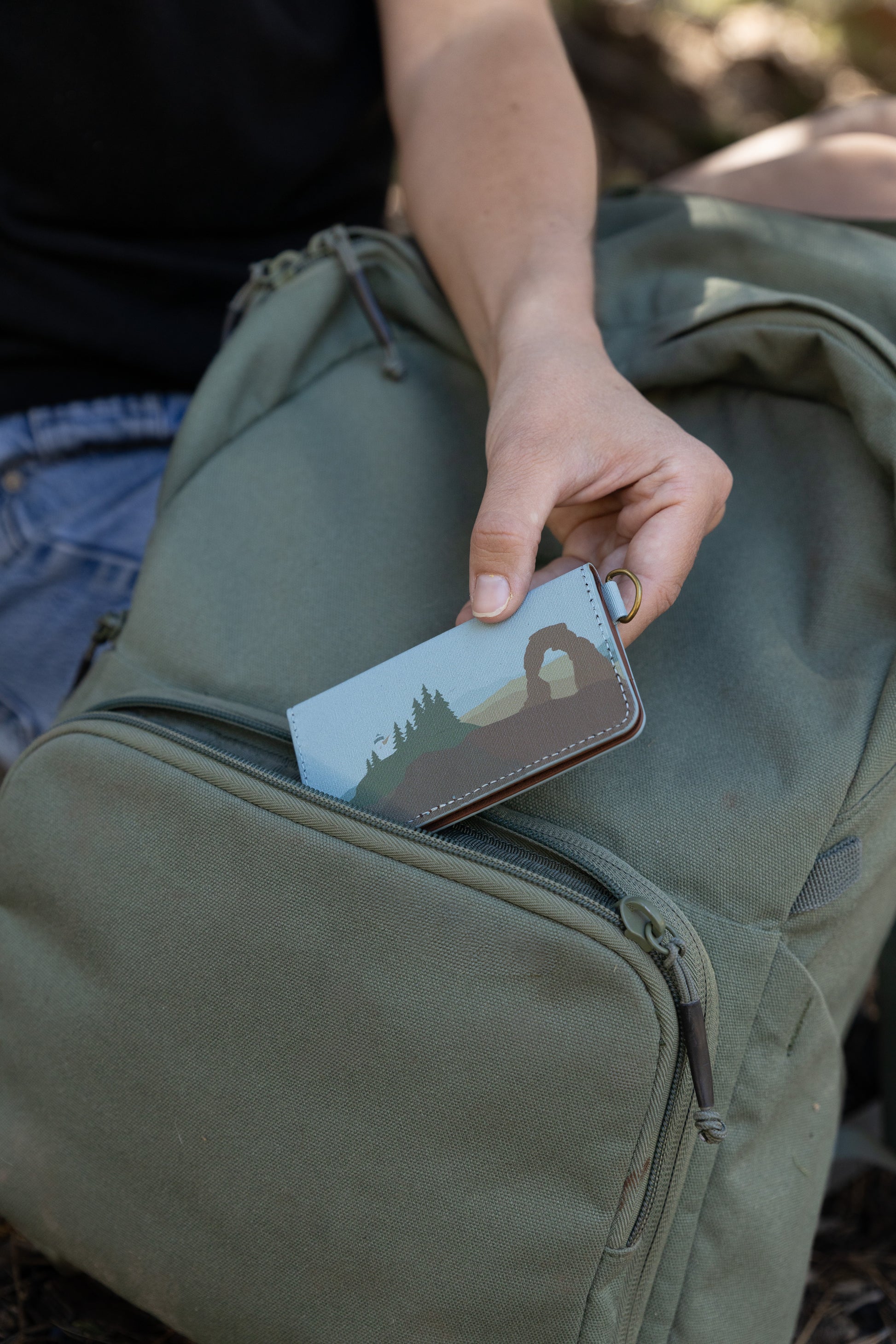 A close-up of a person sliding a Wildtree "Parks in Color" SD card holder into the front pocket of a green canvas backpack. The holder's cover features a brown and green illustration of Delicate Arch and pine trees against a light blue background.