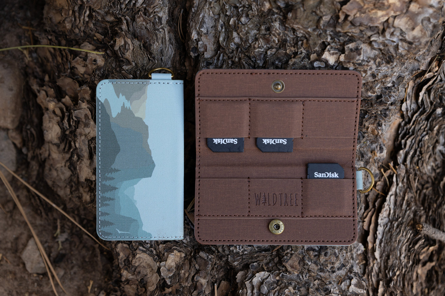 A closed and an open Wildtree "Parks in Color" SD card holder displayed on textured tree bark. The closed wallet features a stylized, layered mountain landscape with pine trees against a pale blue background. The open wallet reveals six brown fabric storage slots, with three SanDisk SD cards peeking out.