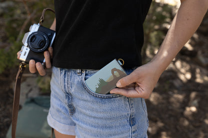 A person holding a Fujifilm camera in one hand while slipping the slim Wildtree "Parks in Color" SD card holder into the front pocket of their denim shorts. The holder features a National Park illustration of Delicate Arch and pine trees.