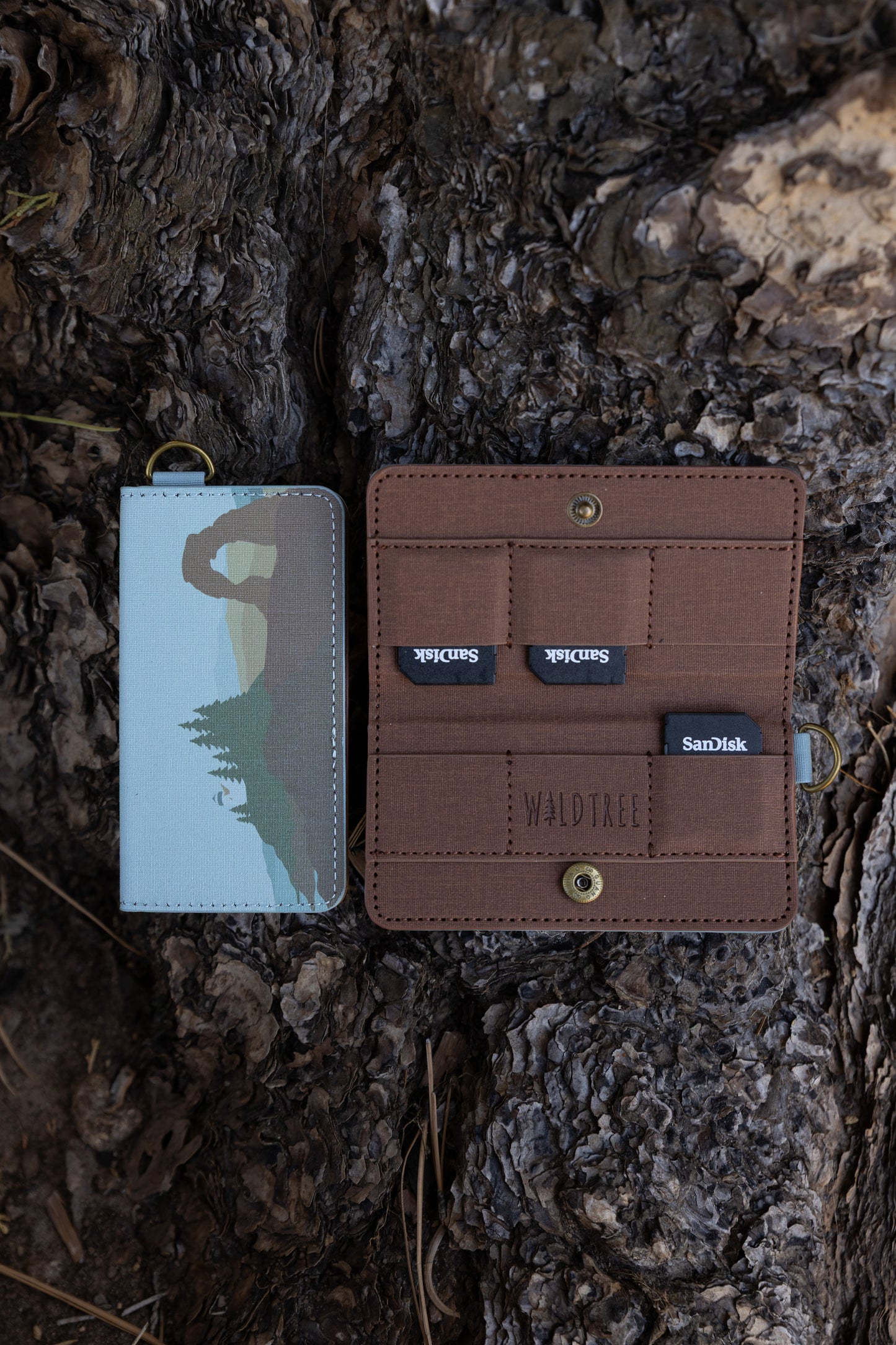 A closed and an open Wildtree "Parks in Color" SD card holder resting on rough tree bark. The closed holder on the left shows an illustrated landscape featuring Delicate Arch, pine trees, and a light blue sky. The open holder on the right shows a brown fabric interior with slots holding three SanDisk SD cards and an embossed Wildtree logo.
