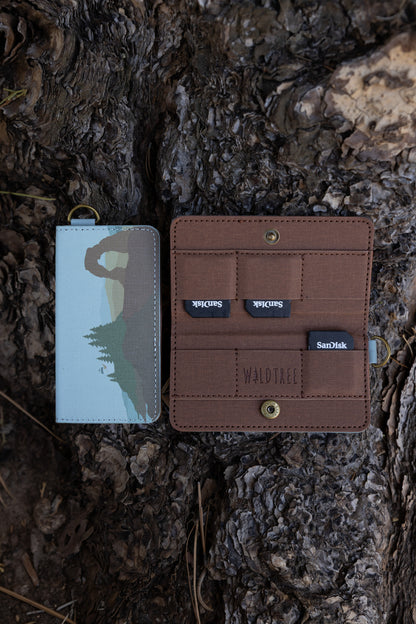 A closed and an open Wildtree "Parks in Color" SD card holder resting on rough tree bark. The closed holder on the left shows an illustrated landscape featuring Delicate Arch, pine trees, and a light blue sky. The open holder on the right shows a brown fabric interior with slots holding three SanDisk SD cards and an embossed Wildtree logo.