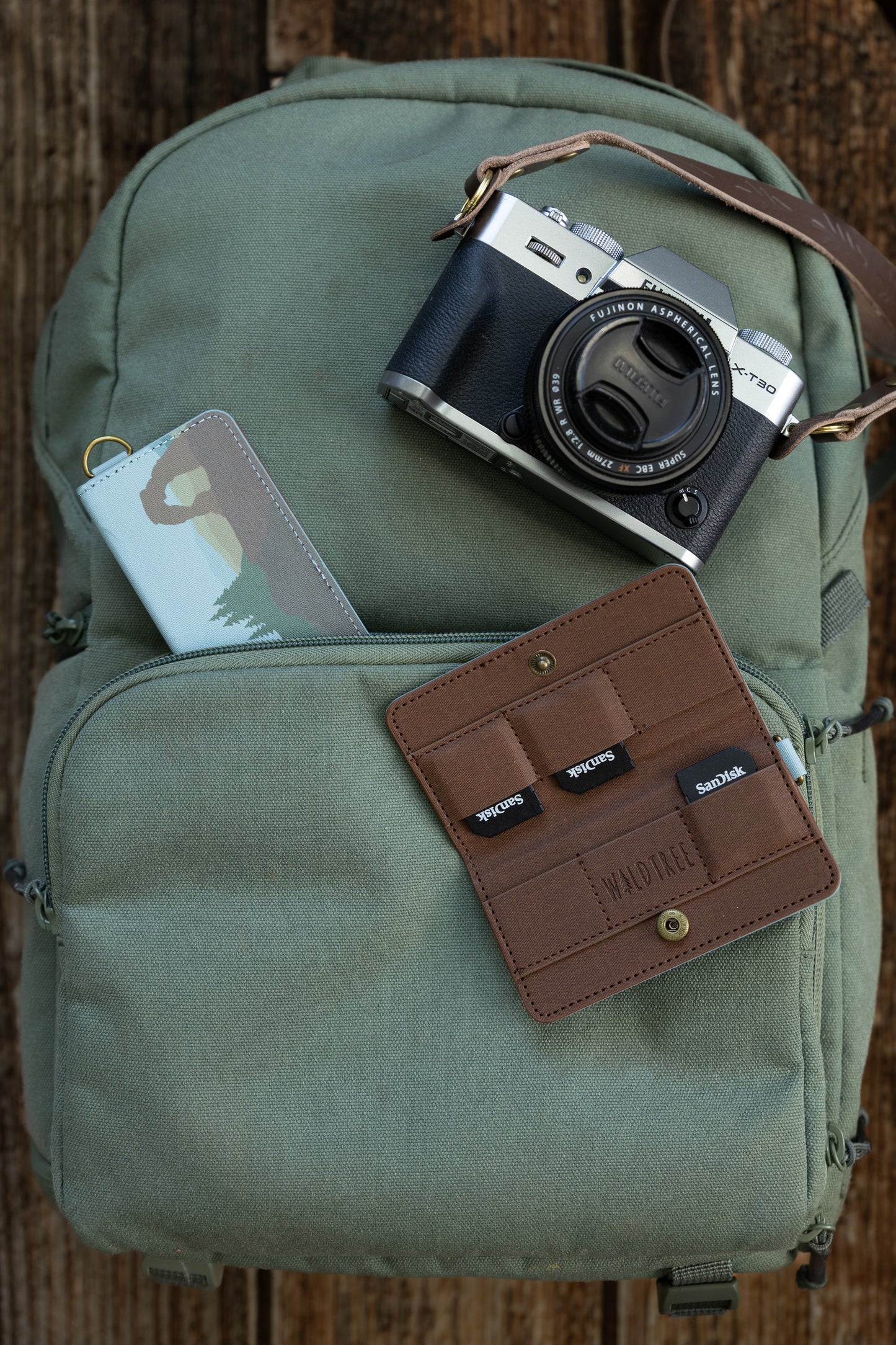 A flat-lay view on a wooden surface showing a green canvas backpack with a silver Fujifilm X-T30 camera on top. A closed "Parks in Color" SD card holder sticks out of the zipper pocket, while an open brown interior holder displaying three SanDisk SD cards lies nearby.