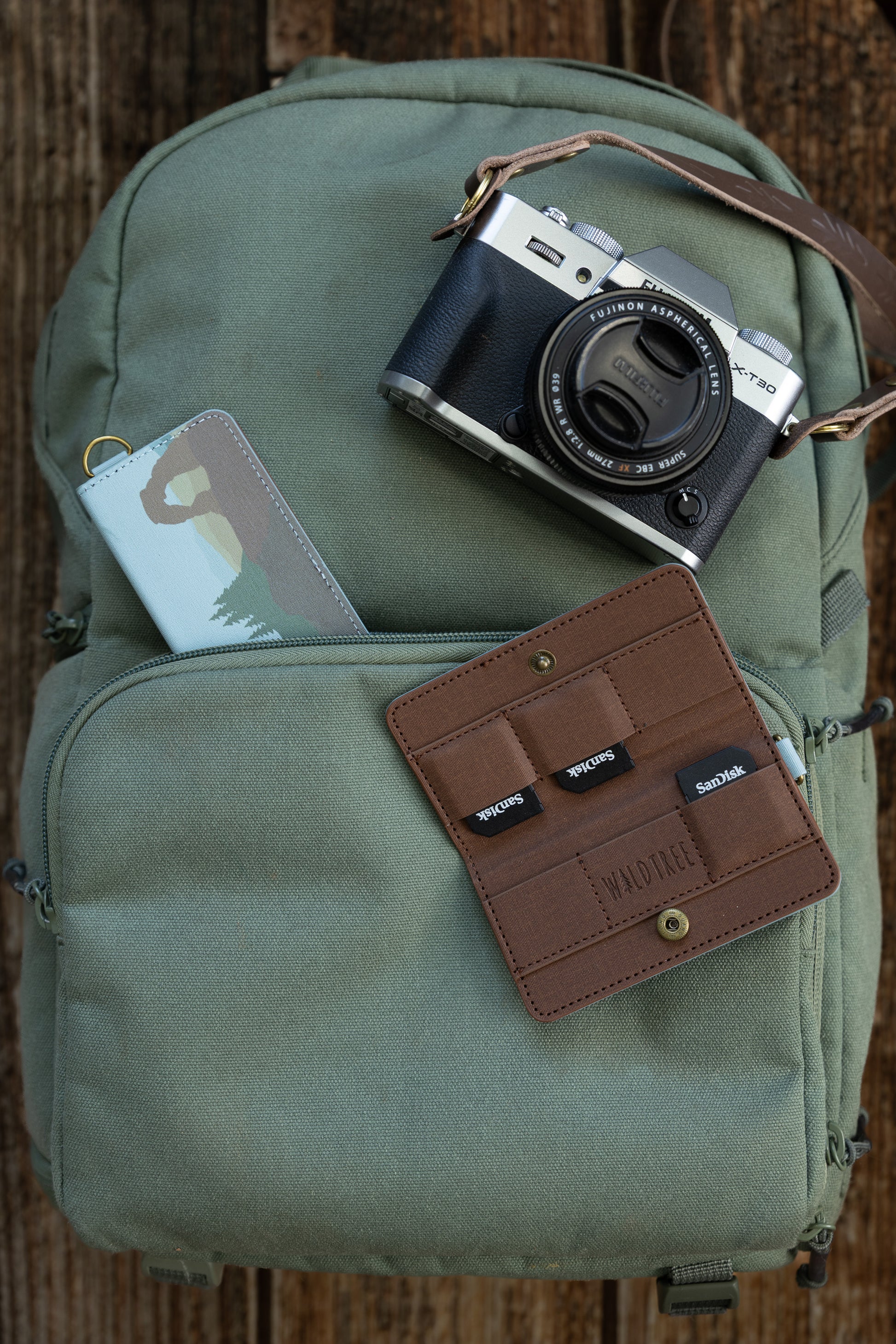 A flat-lay view on a wooden surface showing a green canvas backpack with a silver Fujifilm X-T30 camera on top. A closed "Parks in Color" SD card holder sticks out of the zipper pocket, while an open brown interior holder displaying three SanDisk SD cards lies nearby.