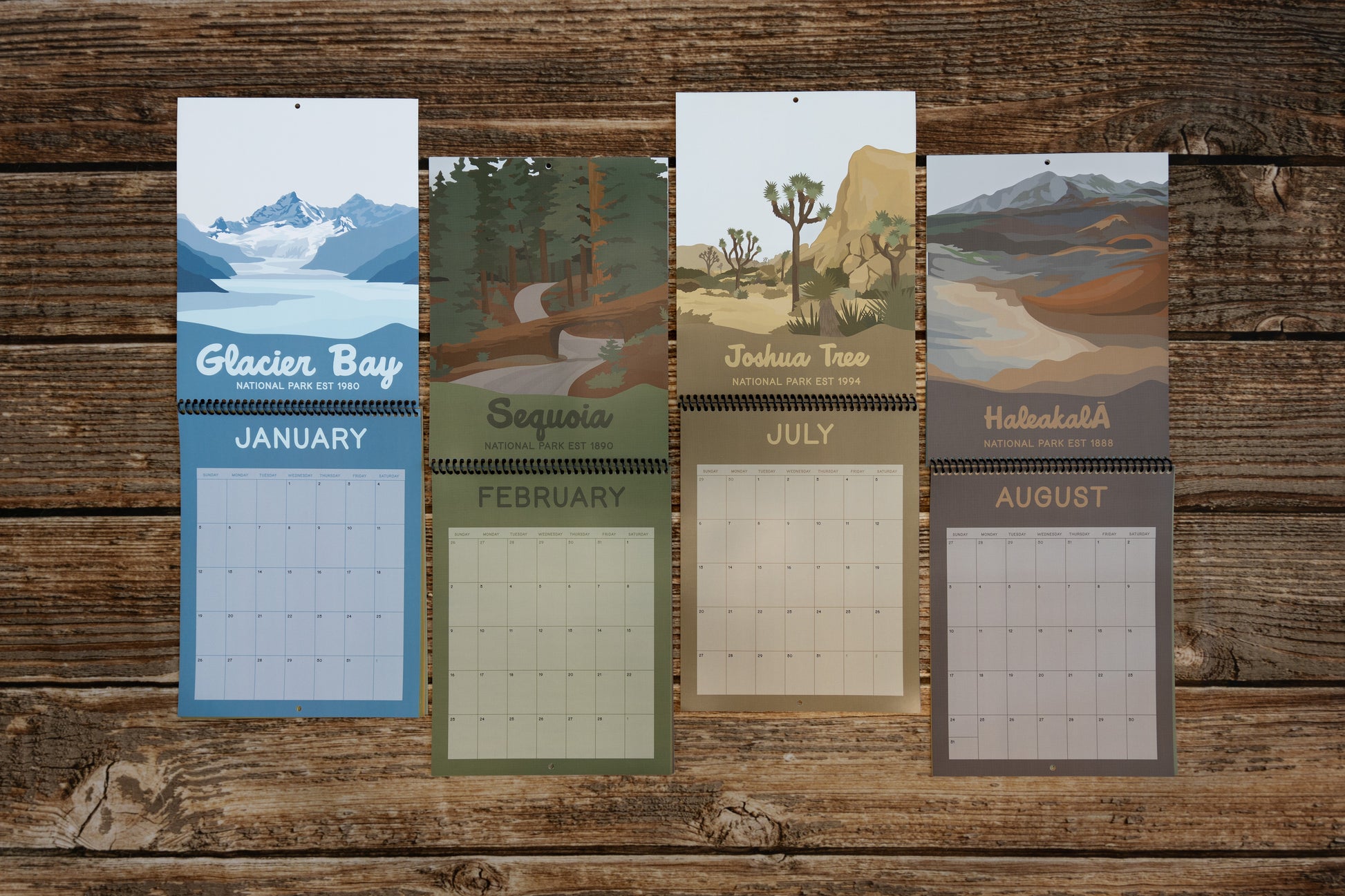 2025 national park calendar. 4 calendars lying on wood floor each showcasing a different month. January - Glacier Bay National park, February - Sequoia National park, July Joshua Tree National park, August - Haleakala National park.