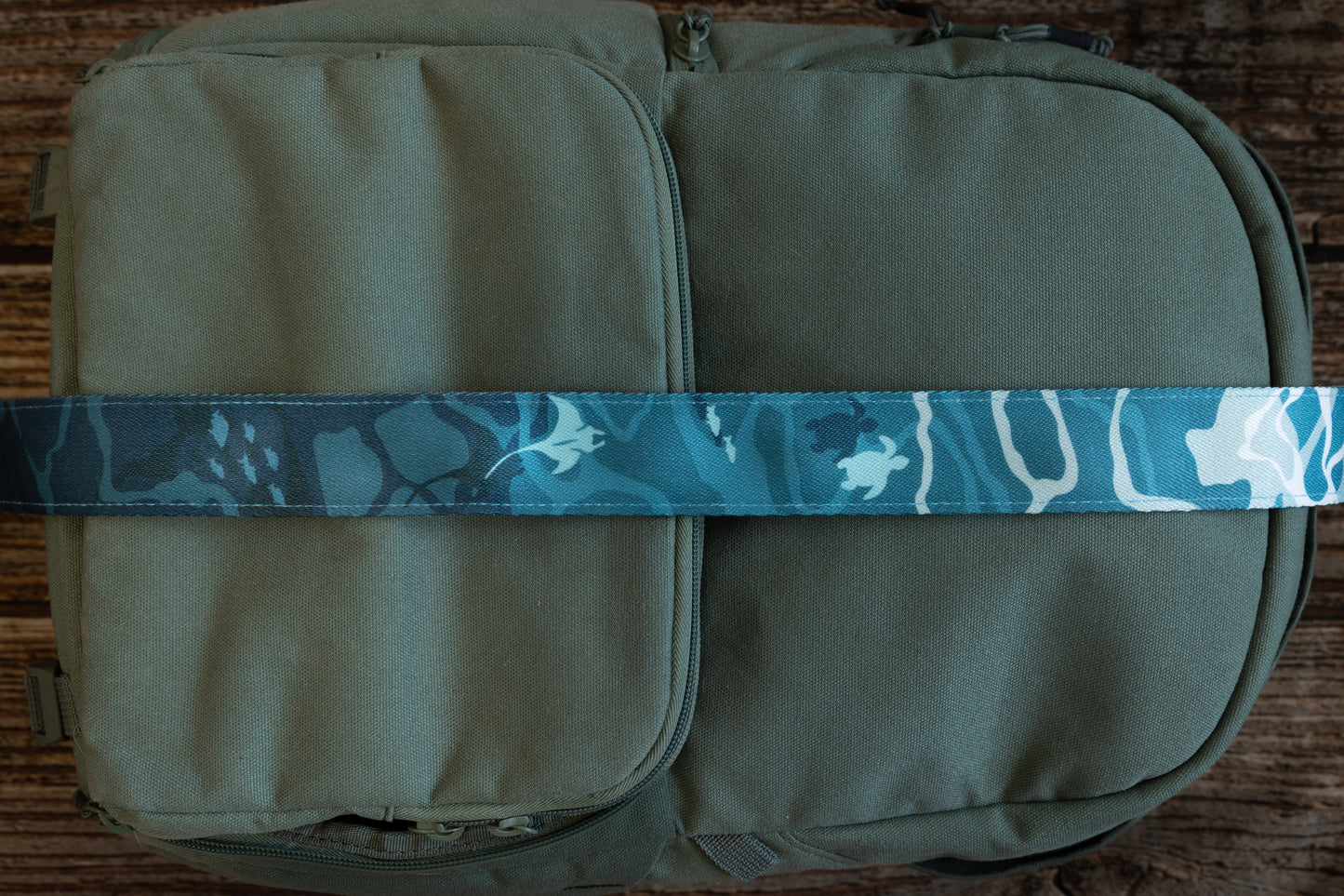 Ocean designed themed Camera Strap. mix of light and dark blues. Ocean water, turtles, sharks, fish and manta rays. light tan leather ends. strap is laying across green back pack