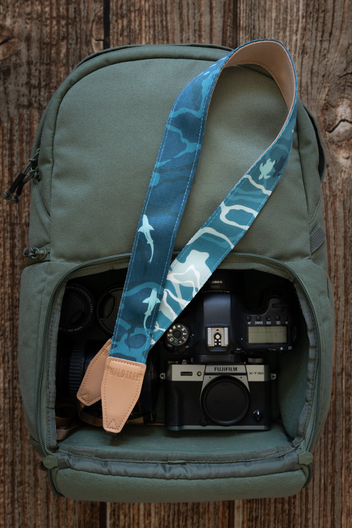 Ocean designed themed Camera Strap. mix of light and dark blues. Ocean water, turtles, sharks, fish and manta rays. light tan leather ends. camera strap is laying out of camera back pack holding cameras