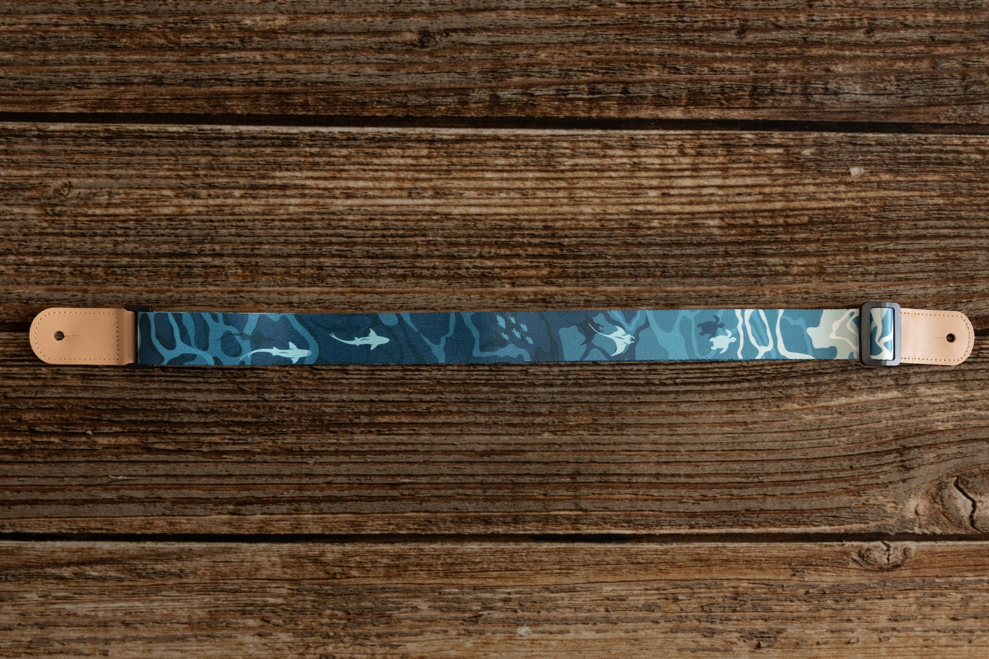 Ocean-designed themed Ukulele Strap. A mix of light and dark blues printed design of ocean water, marine life, turtles, sharks, fish, and manta rays lying on wood floor