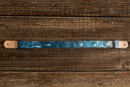 Ocean-designed themed Ukulele Strap. A mix of light and dark blues printed design of ocean water, marine life, turtles, sharks, fish, and manta rays lying on wood floor