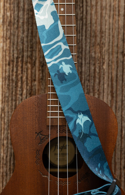 Ocean-designed themed Ukulele Strap. A mix of light and dark blues printed design of ocean water, marine life, turtles, sharks, fish, and manta rays Attached Ukulele lying on wood floor